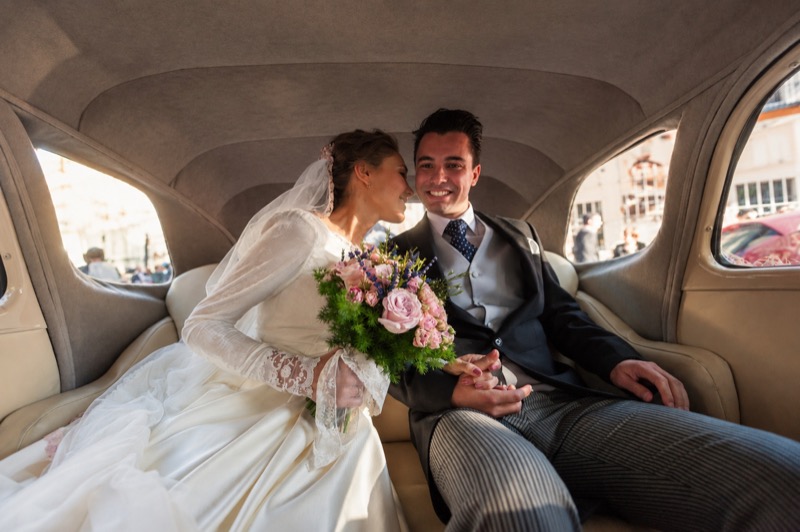 Los novios en el asiento trasero de un coche clasico, con el ramo, camino del banquete
