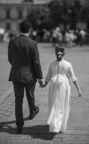 Un padre y su hija de comunion caminan de la mano de espaldas por una plaza empedrada, en blanco y negro
